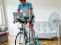 Mężczyzna w stroju kolarskim wykonuje trening wytrzymałościowy na rowerze stacjonarnym w domu, z laptopem przed sobą i wentylatorem obok.