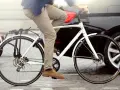 Mężczyzna na rowerze w mieście, obok samochodu i skutera. Rower posiada błotniki i bagażnik, które są obowiązkowym wyposażeniem roweru.