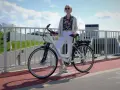 Kobieta w białych spodniach i kolorowej koszuli stoi obok białego roweru elektrycznego. Czy na rower elektryczny trzeba mieć prawo jazdy?
