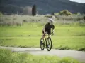Kolarz w kasku i okularach na rowerze, pochylony do przodu, wykonuje intensywny trening beztlenowy na malowniczej drodze.