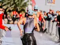 Zawodnicy triathlonu olimpijskiego biegną, a strażak polewa ich wodą z węża, by ich ochłodzić. Widownia w maskach obserwuje zza barierek.
