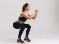 Kobieta w czarnym sportowym stroju wykonuje ćwiczenie air squats, z rękami splecionymi przed sobą.