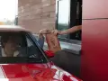 Mężczyzna w czerwonym aucie odbiera z okienka KFC torbę z jedzeniem. Wygląda na zadowolonego, że otrzymał swój mega pocket kcal.