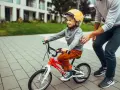 Chłopiec w kasku na czerwonym rowerze 14 cali, idealnym na jego wzrost, uczy się jeździć z pomocą dorosłego.