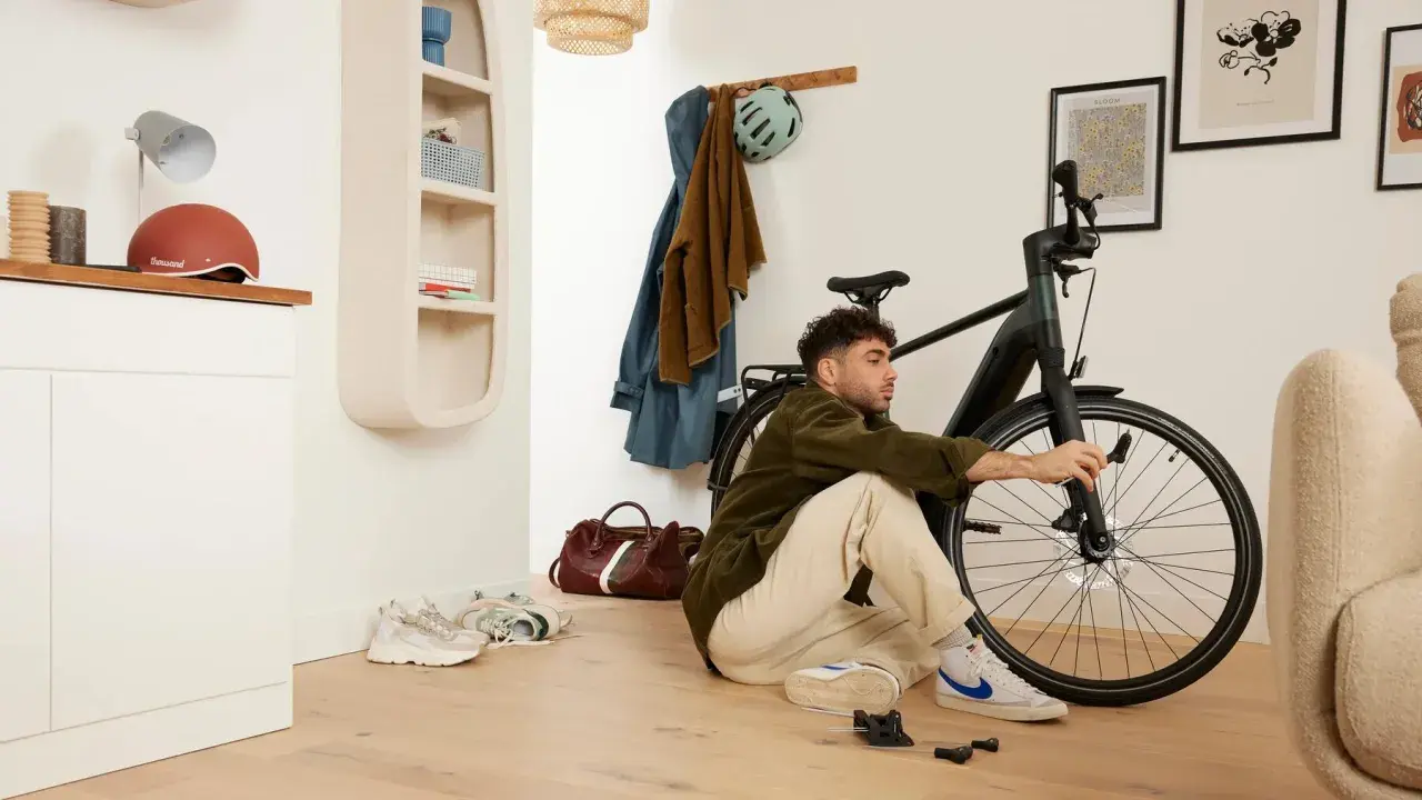 Mężczyzna siedzi na podłodze i majstruje przy rowerze. W tle wieszak z ubraniami i kask, a obok niego **scentrowane koło** roweru.