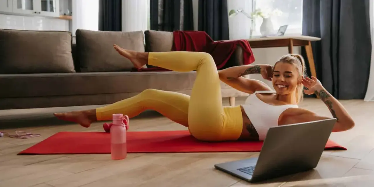 Uśmiechnięta kobieta wykonuje proste ćwiczenia w domu, leżąc na macie i patrząc na laptopa.