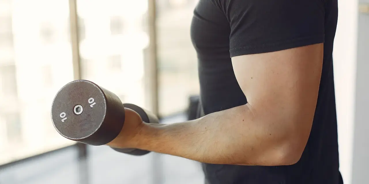 Mężczyzna w czarnej koszulce wykonuje ćwiczenia na biceps z hantlem 10 kg. To jedne z najlepszych ćwiczeń na biceps.