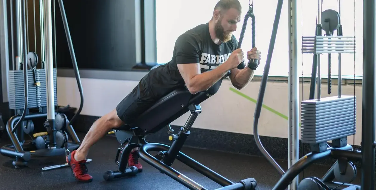 Mężczyzna wykonuje ćwiczenie na triceps, znane jako skull crushers, na ławce skośnej z użyciem wyciągu.