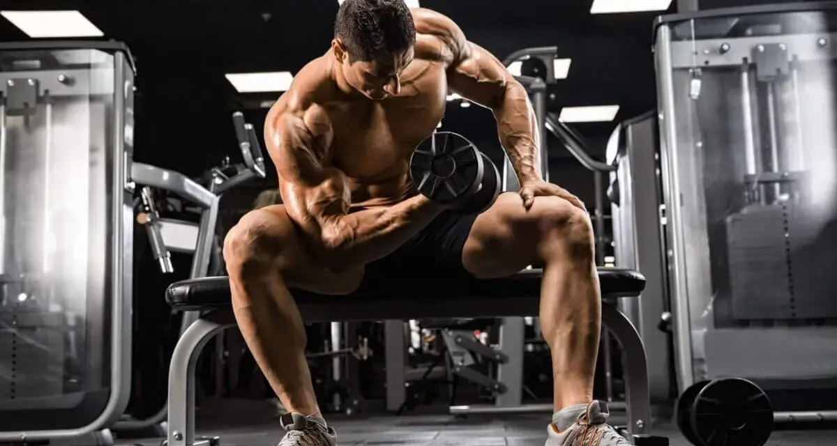 Mężczyzna z wyrzeźbionymi mięśniami wykonuje ćwiczenie na biceps z hantlem, skupiony na swoim **full body workout** w siłowni.