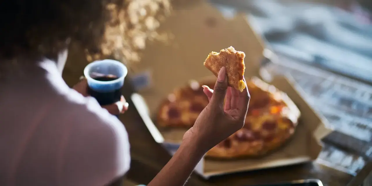 Kobieta trzyma kawałek pizzy i napój, czując głód. Grelina działa, apetyt rośnie.