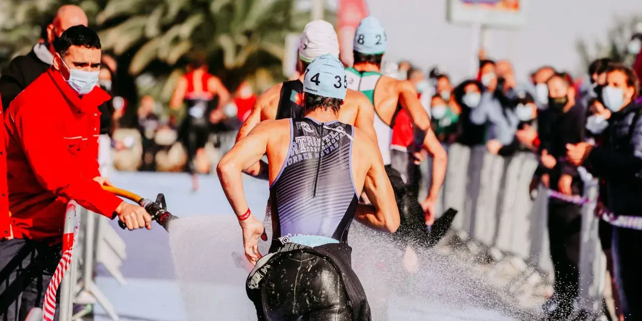 Zawodnicy triathlonu olimpijskiego biegną, a strażak polewa ich wodą z węża, by ich ochłodzić. Widownia w maskach obserwuje zza barierek.