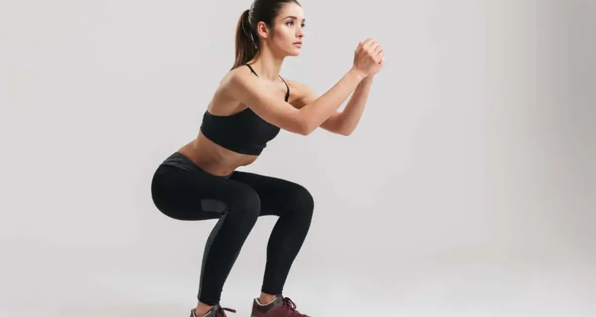 Kobieta w czarnym sportowym stroju wykonuje ćwiczenie air squats, z rękami splecionymi przed sobą.