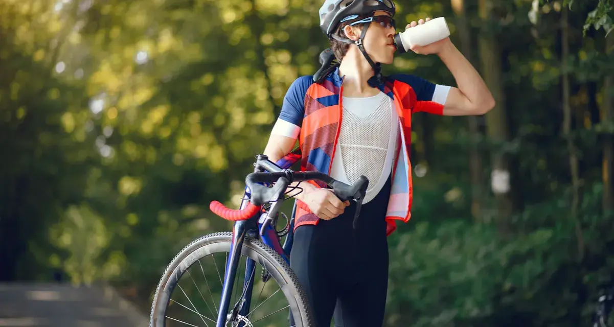 Kolarz w kasku i stroju sportowym pije wodę, trzymając rower. Dba o nawodnienie i dostarczenie makroskładników po wysiłku.
