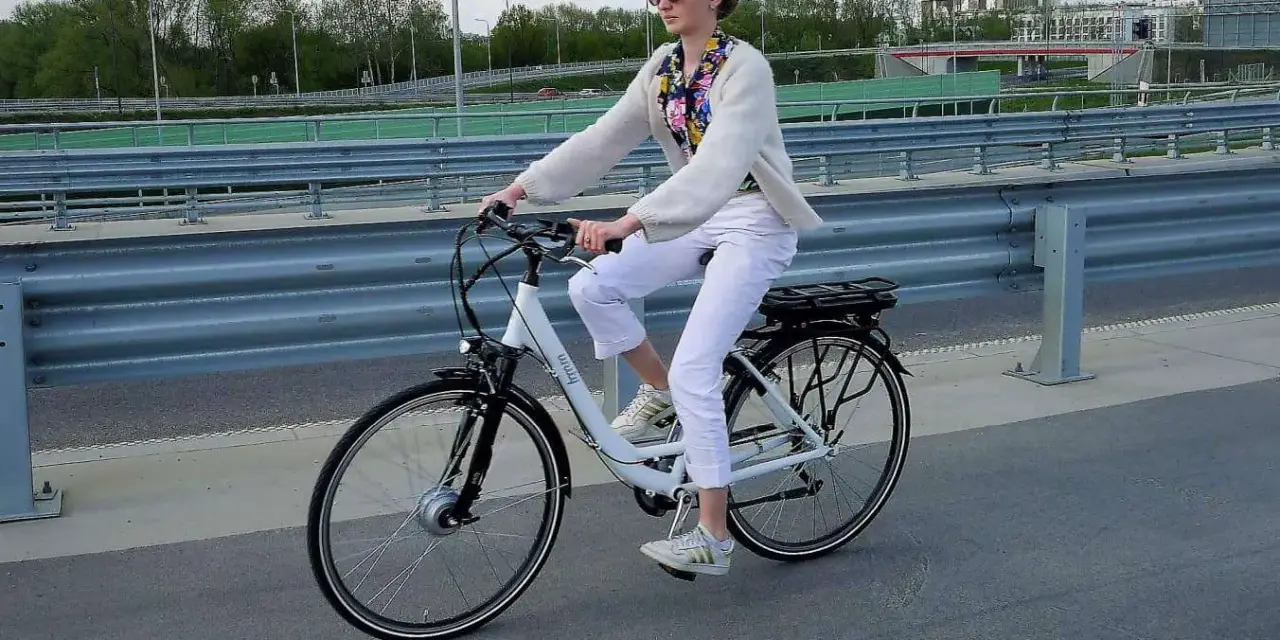 Kobieta w białych spodniach i sweterku jedzie na rowerze elektrycznym. Przepisy dotyczące rowerów elektrycznych są ważne dla bezpieczeństwa.
