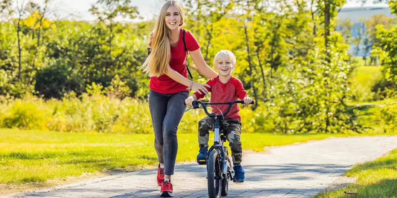 Mama uczy synka, jak nauczyć dziecko jeździć na rowerze. Radosny chłopiec pedałuje, a mama go asekuruje.