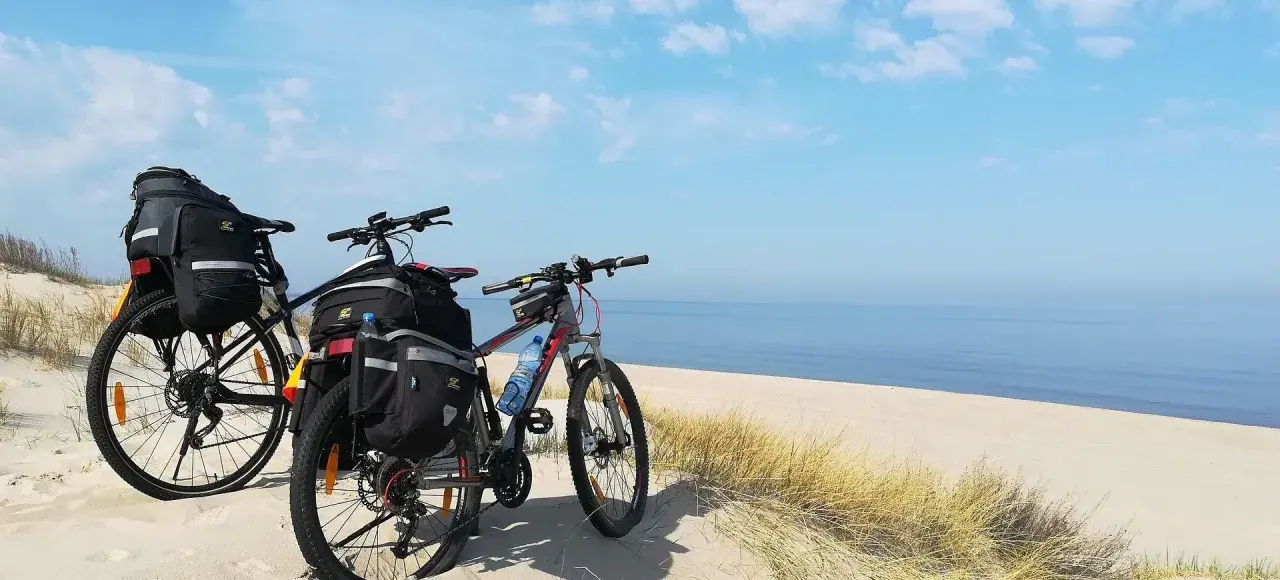 Dwa rowery z sakwami na plaży, gotowe na przygodę wzdłuż Velo Baltica. Morze i niebo tworzą tło.