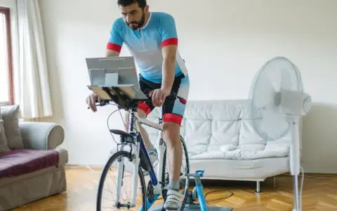 Mężczyzna w stroju kolarskim wykonuje trening wytrzymałościowy na rowerze stacjonarnym w domu, z laptopem przed sobą i wentylatorem obok.