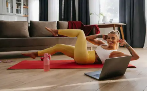 Uśmiechnięta kobieta wykonuje proste ćwiczenia w domu, leżąc na macie i patrząc na laptopa.