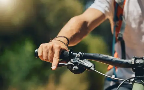 Mężczyzna trzyma kierownicę roweru, jego przedramię jest lekko spięte. Słońce przebija się przez liście.