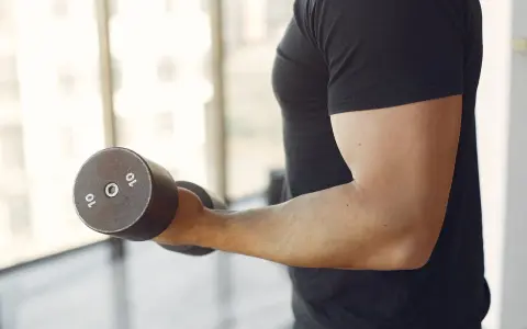 Mężczyzna w czarnej koszulce wykonuje ćwiczenia na biceps z hantlem 10 kg. To jedne z najlepszych ćwiczeń na biceps.