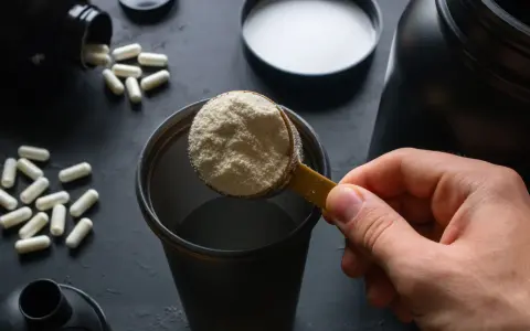 Łyżka białego proszku, czyli protein, trafia do shakera. Obok leżą kapsułki.