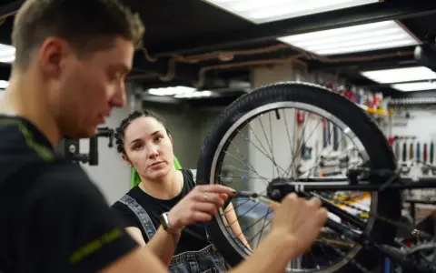 Mechanik rowerowy wyjaśnia, ile kosztuje wymiana dętki w rowerze, podczas gdy kobieta z zielonymi włosami uważnie słucha.