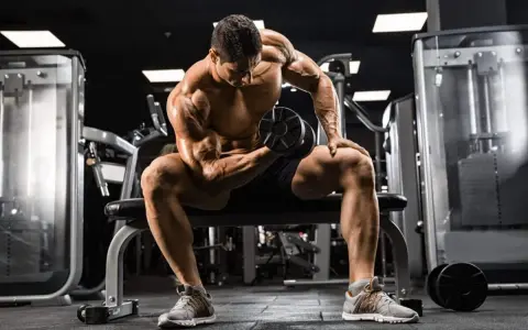 Mężczyzna z wyrzeźbionymi mięśniami wykonuje ćwiczenie na biceps z hantlem, skupiony na swoim **full body workout** w siłowni.