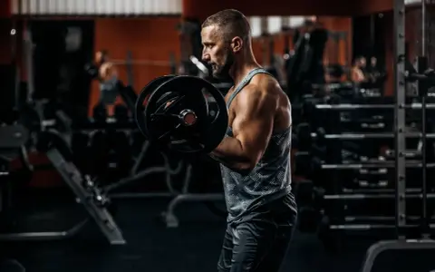 Mężczyzna ćwiczy biceps z ciężarem. W tle siłownia, sprzęt i inny ćwiczący.