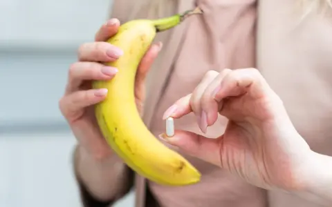 Kobieta trzyma banana i tabletkę, zastanawiając się, ile potasu dziennie powinna przyjmować.