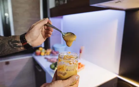 Mężczyzna z tatuażem na ramieniu nabiera łyżką gęsty sos z słoika. Czy musztarda jest zdrowa? W tle widać kuchnię i warzywa.