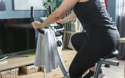 Kobieta ćwiczy na rowerku stacjonarnym, osiągając efekty w domowym zaciszu. Obok ręcznik i telewizor.