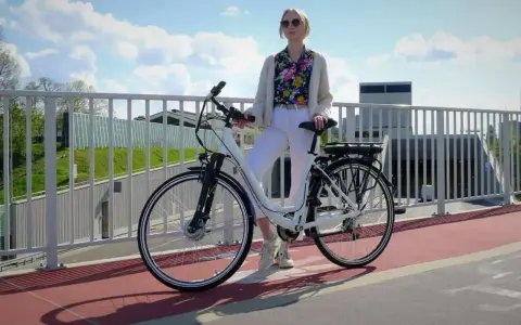 Kobieta w białych spodniach i kolorowej koszuli stoi obok białego roweru elektrycznego. Czy na rower elektryczny trzeba mieć prawo jazdy?