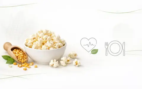 Misternie przygotowany popcorn w misce, obok ziaren kukurydzy na drewnianej łyżce. Czy popcorn tuczy? Ikony serca i talerza sugerują zdrowy wybór.