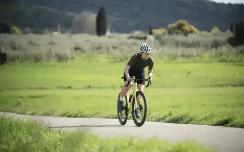 Kolarz w kasku i okularach na rowerze, pochylony do przodu, wykonuje intensywny trening beztlenowy na malowniczej drodze.
