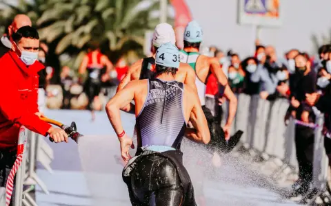 Zawodnicy triathlonu olimpijskiego biegną, a strażak polewa ich wodą z węża, by ich ochłodzić. Widownia w maskach obserwuje zza barierek.