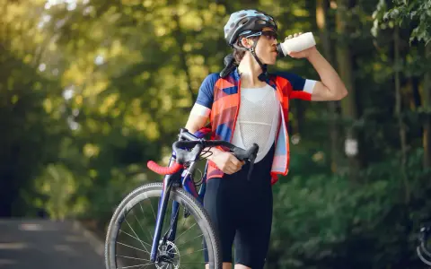 Kolarz w kasku i stroju sportowym pije wodę, trzymając rower. Dba o nawodnienie i dostarczenie makroskładników po wysiłku.