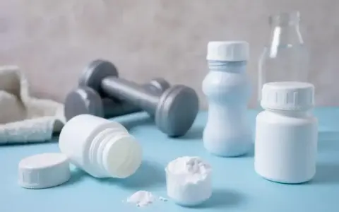 Białe pojemniki, miarka z proszkiem i hantle sugerują suplementację. Proteiny co to? To budulec mięśni, ważny dla sportowców.