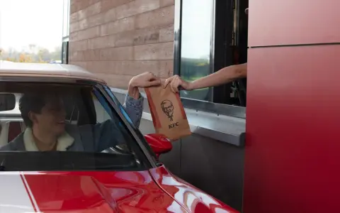 Mężczyzna w czerwonym aucie odbiera z okienka KFC torbę z jedzeniem. Wygląda na zadowolonego, że otrzymał swój mega pocket kcal.