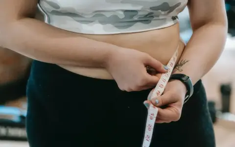 Kobieta mierzy swój duży brzuch centymetrem krawieckim, ubrana w sportowy top i czarne legginsy.