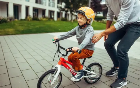 Chłopiec w kasku na czerwonym rowerze 14 cali, idealnym na jego wzrost, uczy się jeździć z pomocą dorosłego.