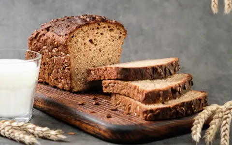 Chleb proteinowy z ziarnami, pokrojony na kromki, obok szklanka mleka i kłosy zboża. Idealny na zdrowe śniadanie.