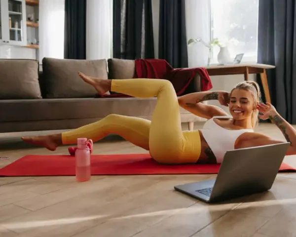 Uśmiechnięta kobieta wykonuje proste ćwiczenia w domu, leżąc na macie i patrząc na laptopa.