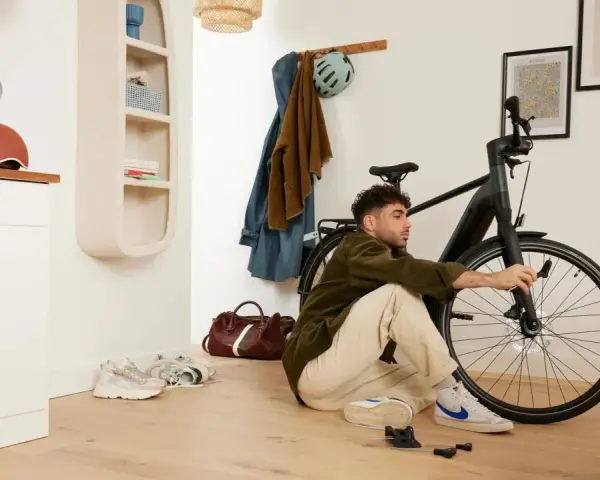 Mężczyzna siedzi na podłodze i majstruje przy rowerze. W tle wieszak z ubraniami i kask, a obok niego **scentrowane koło** roweru.