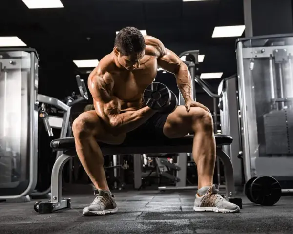 Mężczyzna z wyrzeźbionymi mięśniami wykonuje ćwiczenie na biceps z hantlem, skupiony na swoim **full body workout** w siłowni.
