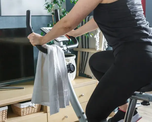 Kobieta ćwiczy na rowerku stacjonarnym, osiągając efekty w domowym zaciszu. Obok ręcznik i telewizor.