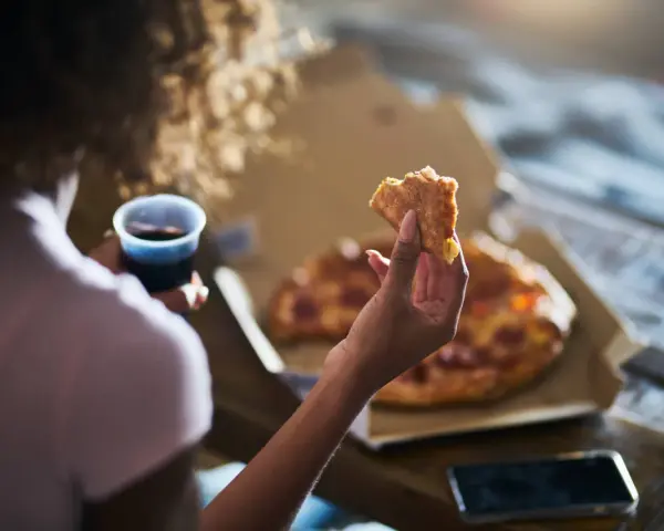 Kobieta trzyma kawałek pizzy i napój, czując głód. Grelina działa, apetyt rośnie.