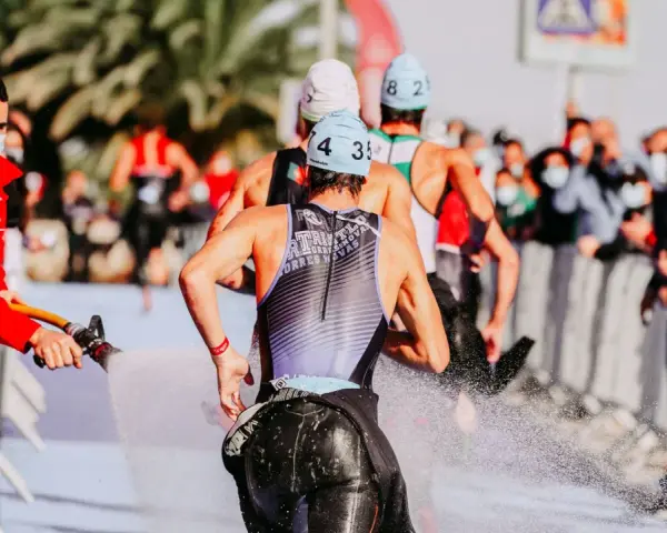 Zawodnicy triathlonu olimpijskiego biegną, a strażak polewa ich wodą z węża, by ich ochłodzić. Widownia w maskach obserwuje zza barierek.