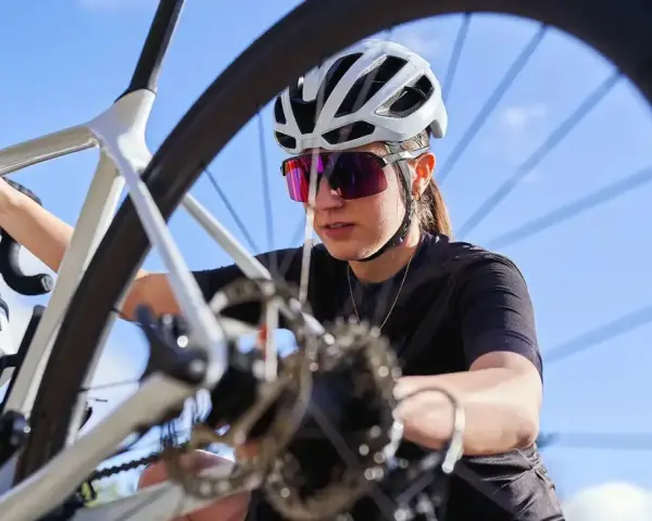 Kobieta w kasku i okularach rowerowych sprawdza rower. Widoczny jest fragment ramy, kierownicy i koła, a także przerzutka.