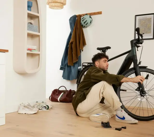 Mężczyzna siedzi na podłodze i majstruje przy rowerze. W tle wieszak z ubraniami i kask, a obok niego **scentrowane koło** roweru.