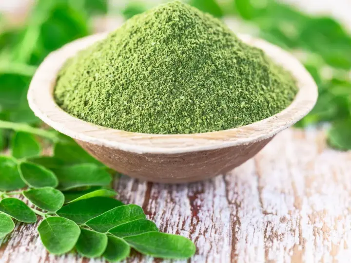 Zielony proszek z liści moringa w drewnianej miseczce, otoczony świeżymi liśćmi. Czy to ściema? Nie, to superfood!